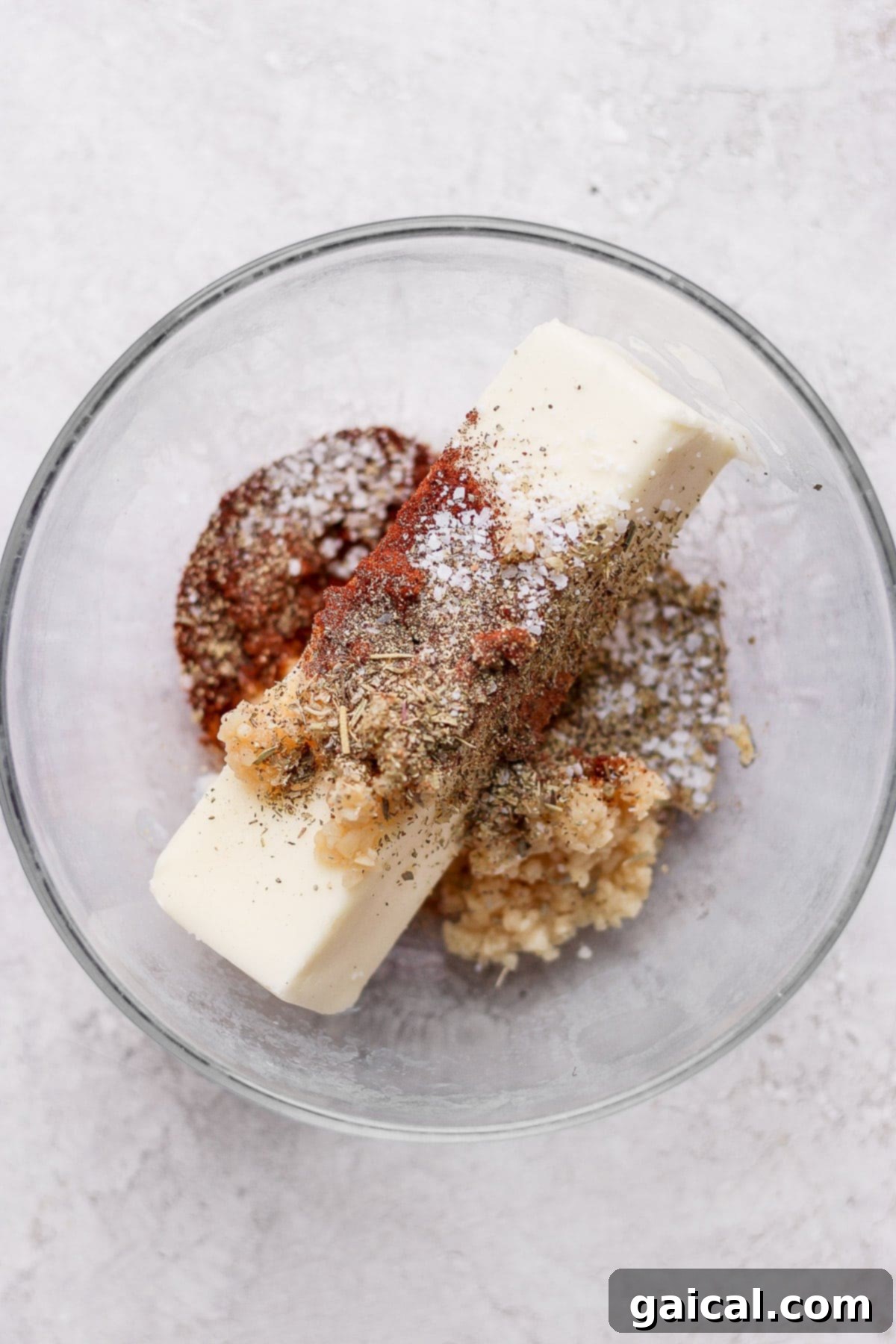 A glass bowl containing softened butter mixed with various colorful spices, ready to be applied to a chicken.