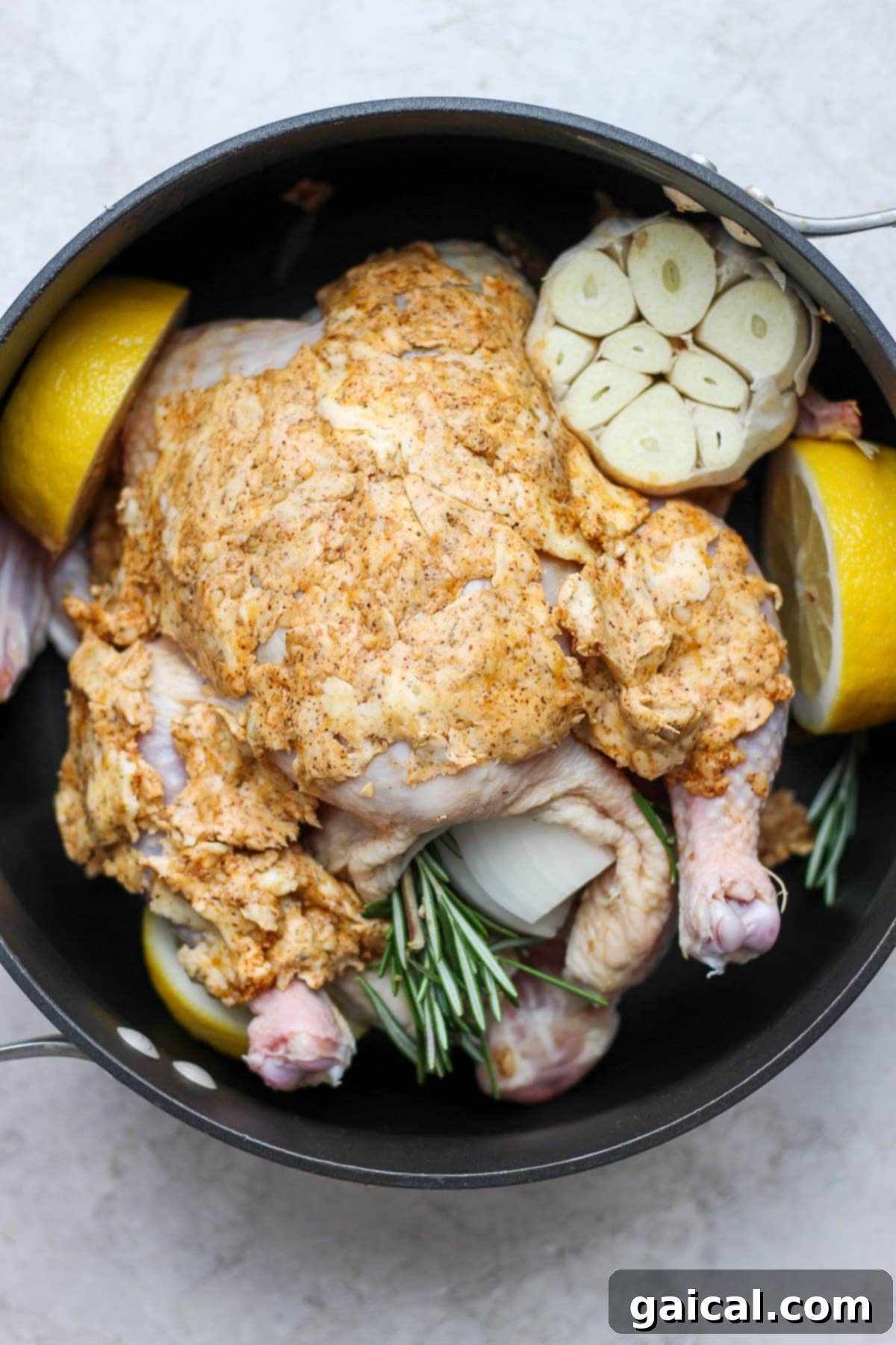 A raw whole chicken, seasoned and ready for roasting, placed in a dark ceramic pan with fresh herbs and lemon slices.