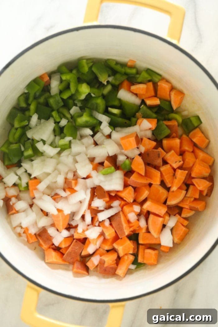 Assorted fresh vegetables like minced yellow onion, diced bell peppers, cubed sweet potatoes, and chopped jalapeños prepped in a large stockpot, ready for cooking.