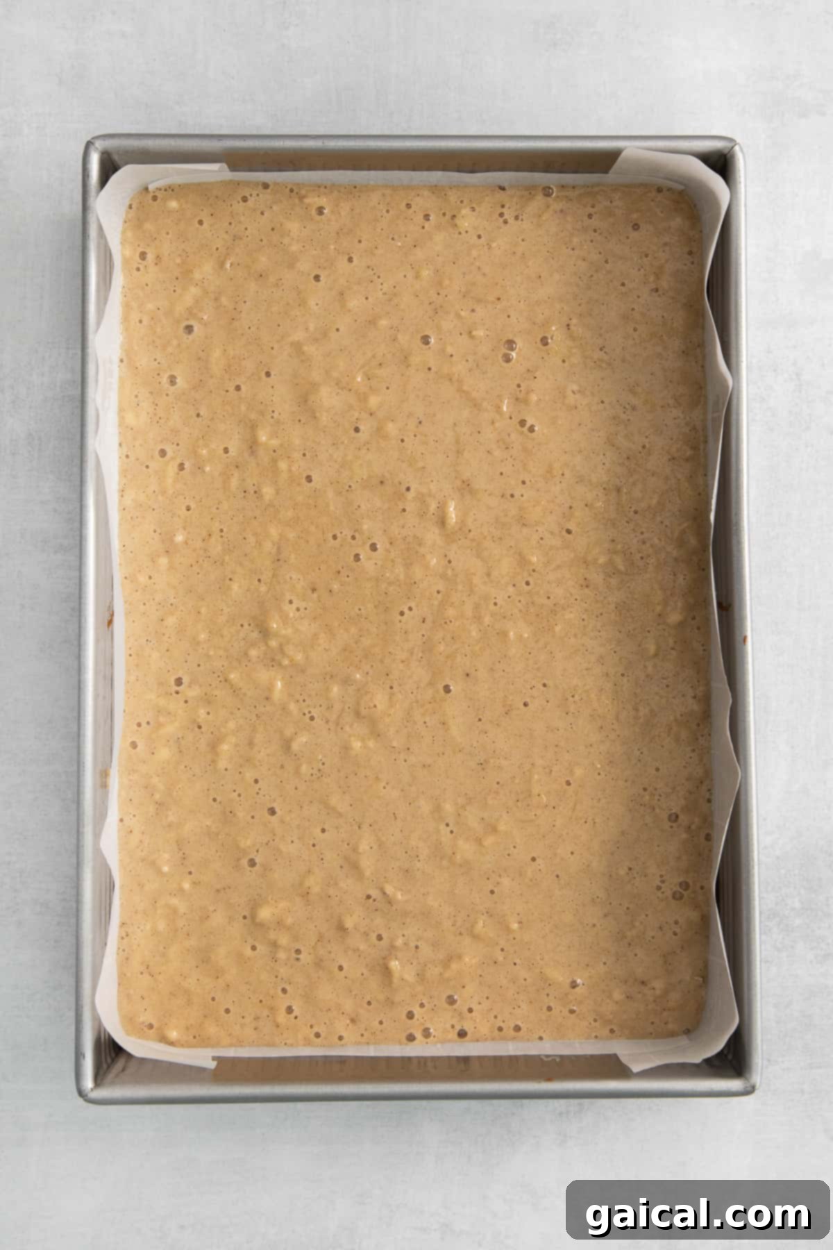 Smooth apple cake batter being carefully poured into a 9x13 inch baking pan, which is lined with parchment paper.