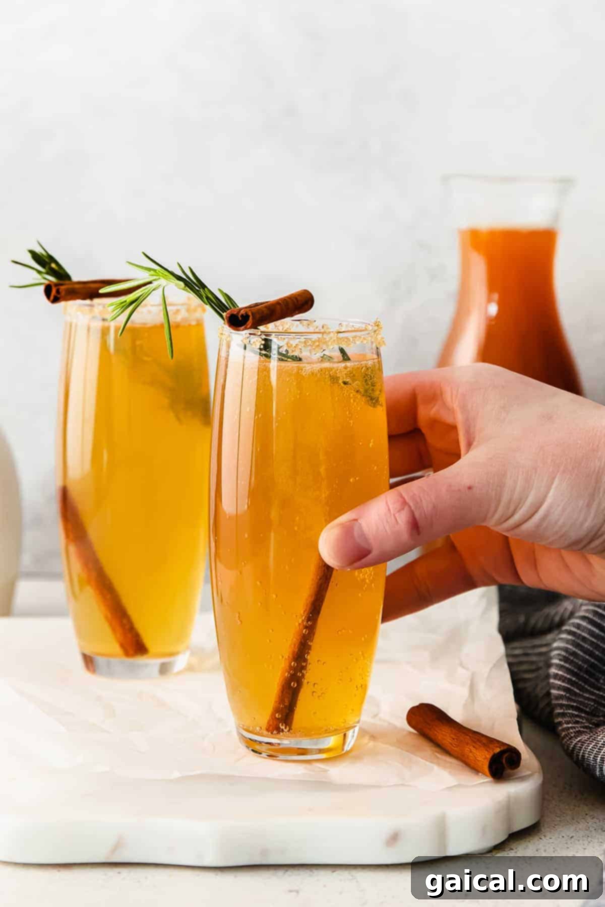 Spiced Apple Mimosa 2 A hand holding an elegant apple cider mimosa cocktail garnished with a rosemary sprig and apple slice, perfect for holiday celebrations.
