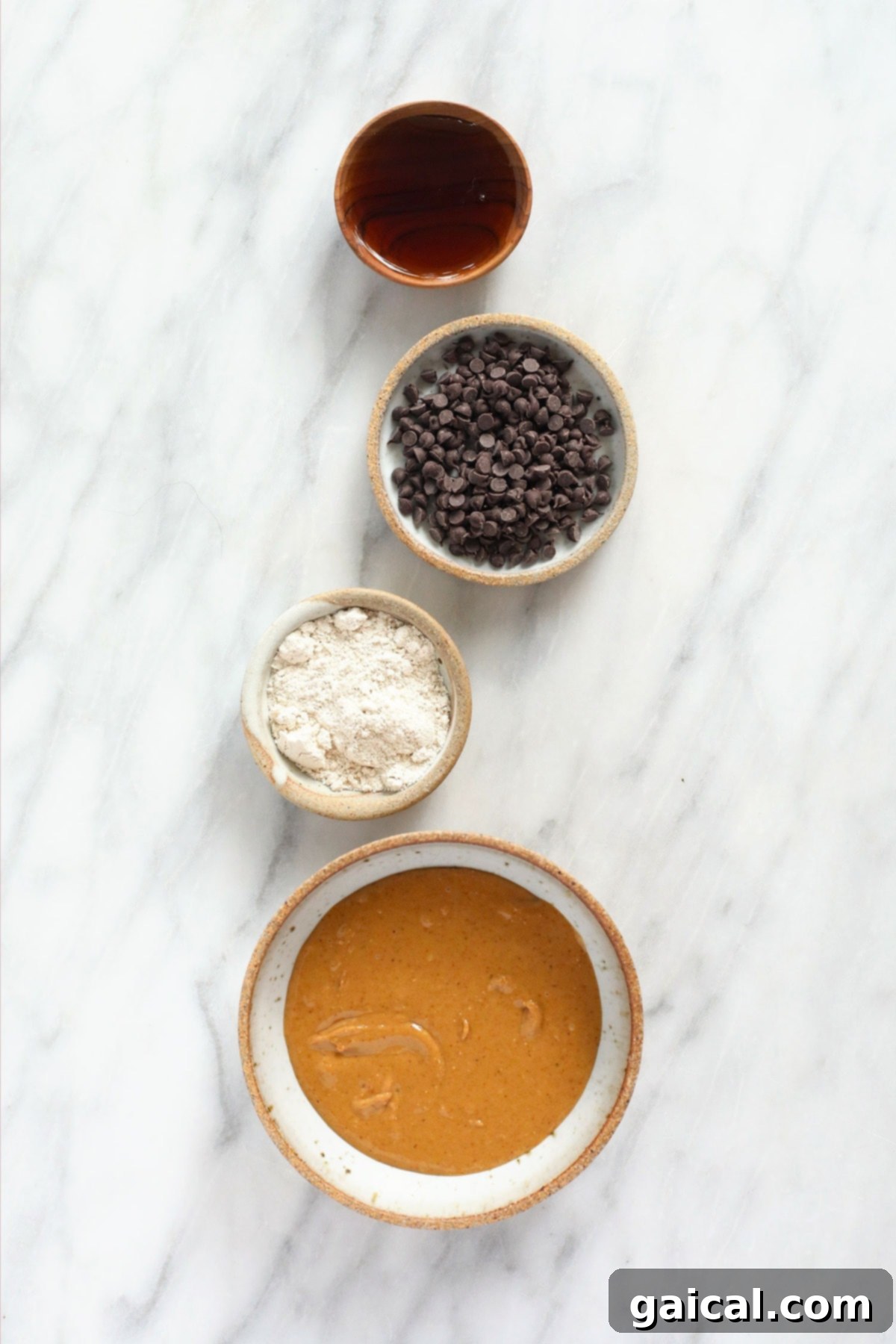 Simple, wholesome ingredients for your homemade vegan cookie dough. Bowls of raw ingredients for vegan cookie dough: maple syrup, dairy-free chocolate chips, oat flour, and creamy peanut butter.