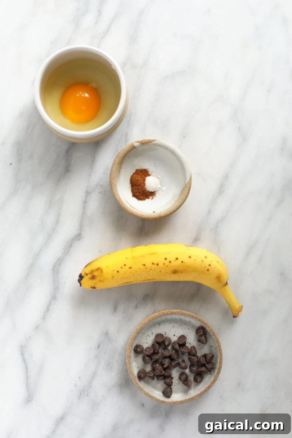 Ingredients for banana mug cake laid out: mashed banana, egg, melted butter, flour, baking powder, cinnamon, coconut sugar, and chocolate chips.