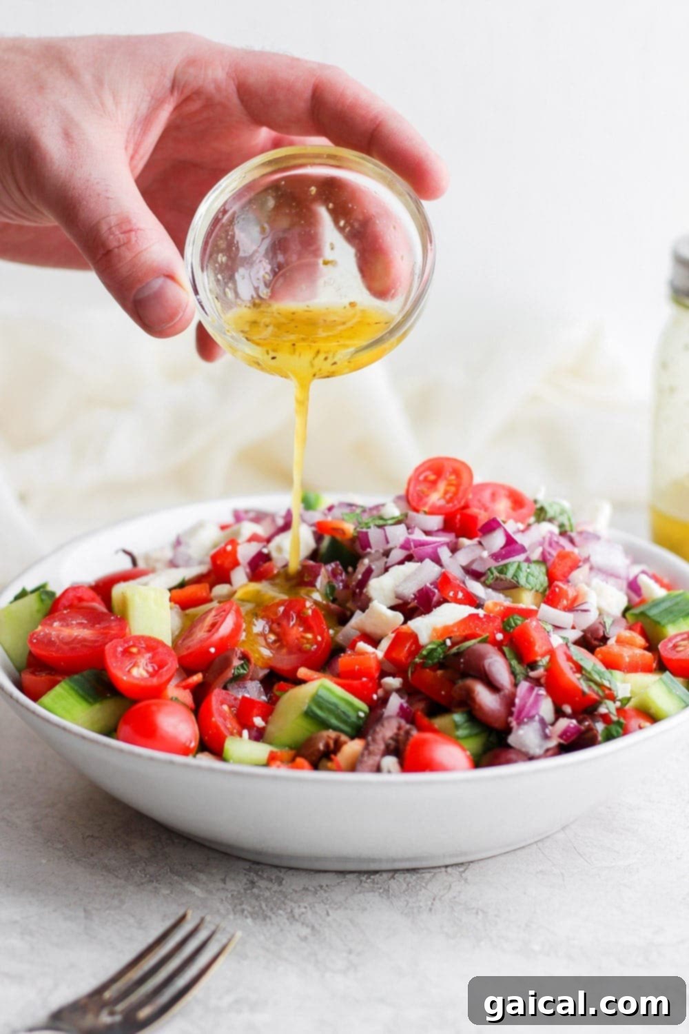 A hand pouring homemade Greek salad dressing over a freshly prepared Greek salad in a large bowl, illustrating the final, delightful step of preparation before serving.