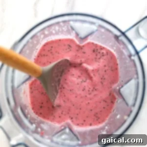 A pink strawberry smoothie in a blender, with a wooden spoon inserted, showing its thick and creamy consistency.