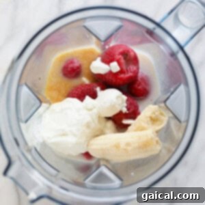 A blender bowl filled with fresh bananas, frozen raspberries, and a dollop of whipped cream, ready to be blended.