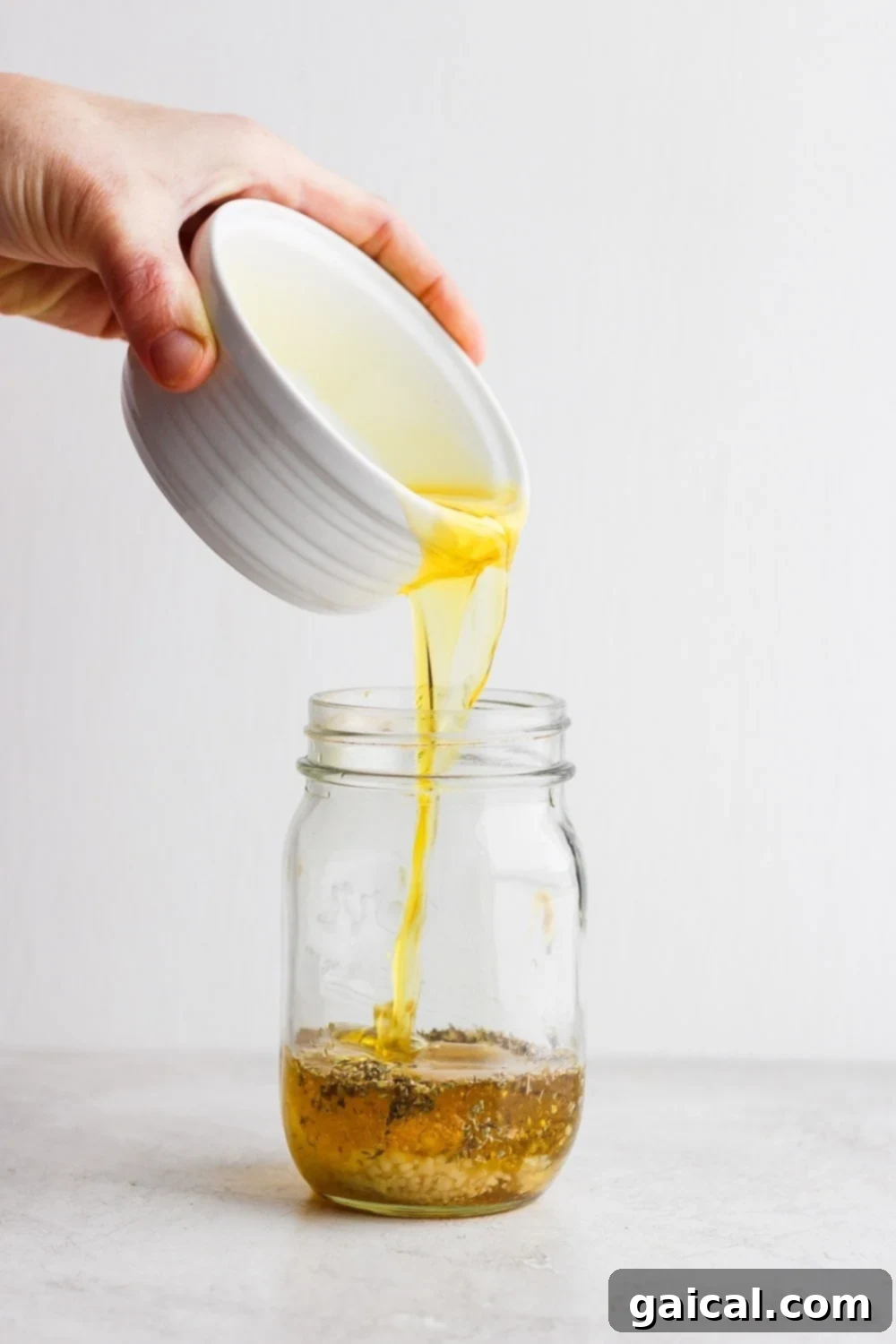 Pouring rich, golden olive oil into a clear glass mason jar, signifying the first step in creating a homemade Greek salad dressing. The oil glistens as it fills the bottom of the jar.