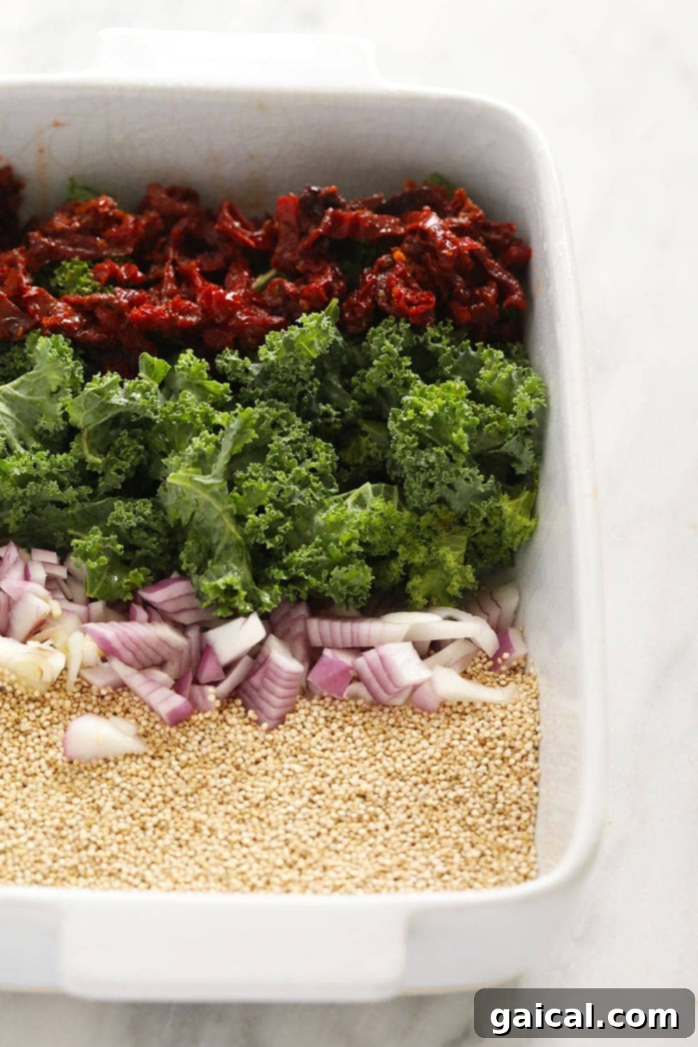 A baking dish filled with uncooked Italian ingredients, including sliced chicken sausage, quinoa, kale, and sun-dried tomatoes, before baking.