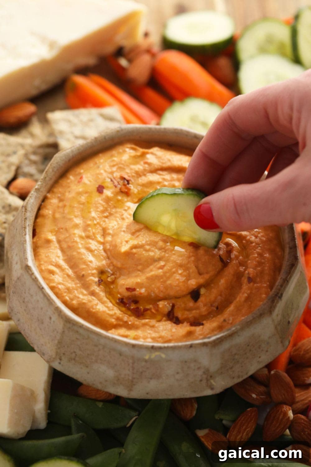Roasted red pepper hummus in a bowl with a cucumber stick dipping into it, showing creamy texture.