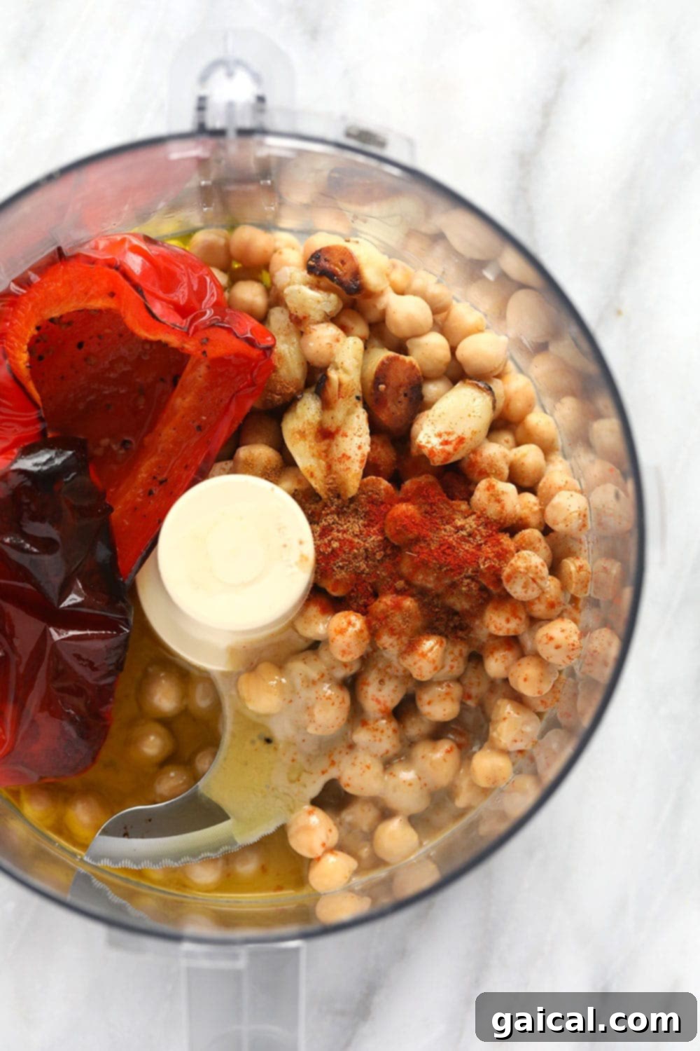 All hummus ingredients, including roasted red peppers, chickpeas, tahini, and lemon juice, are loaded into a food processor and ready for blending.
