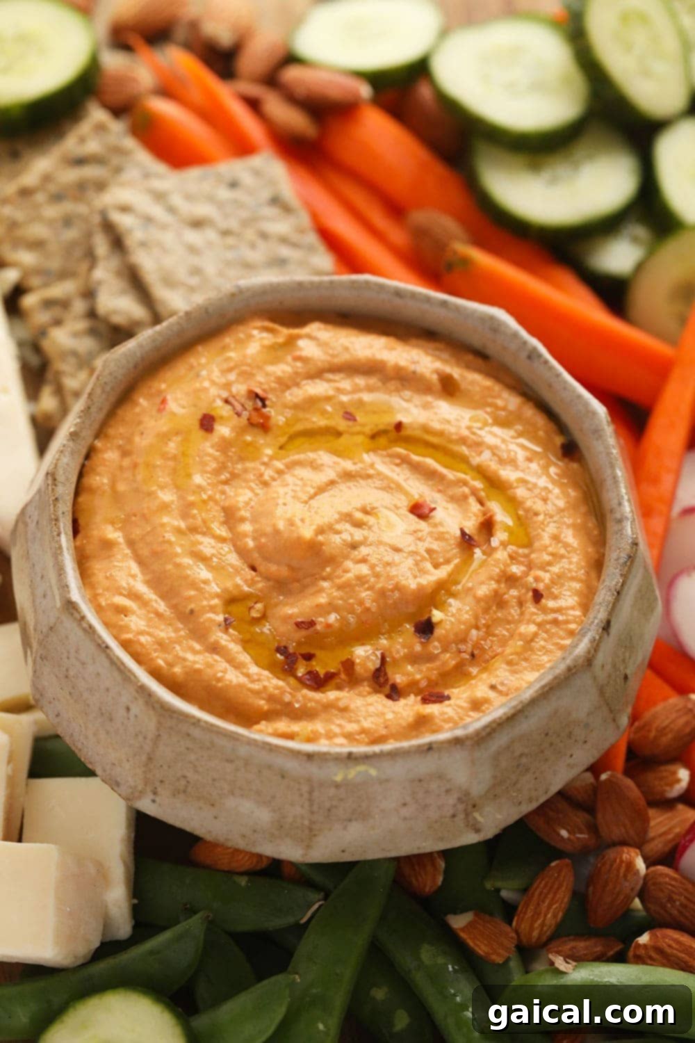 A bowl of vibrant roasted red pepper hummus surrounded by an assortment of fresh vegetables like cucumber and bell pepper slices, and various crackers, ideal for dipping.
