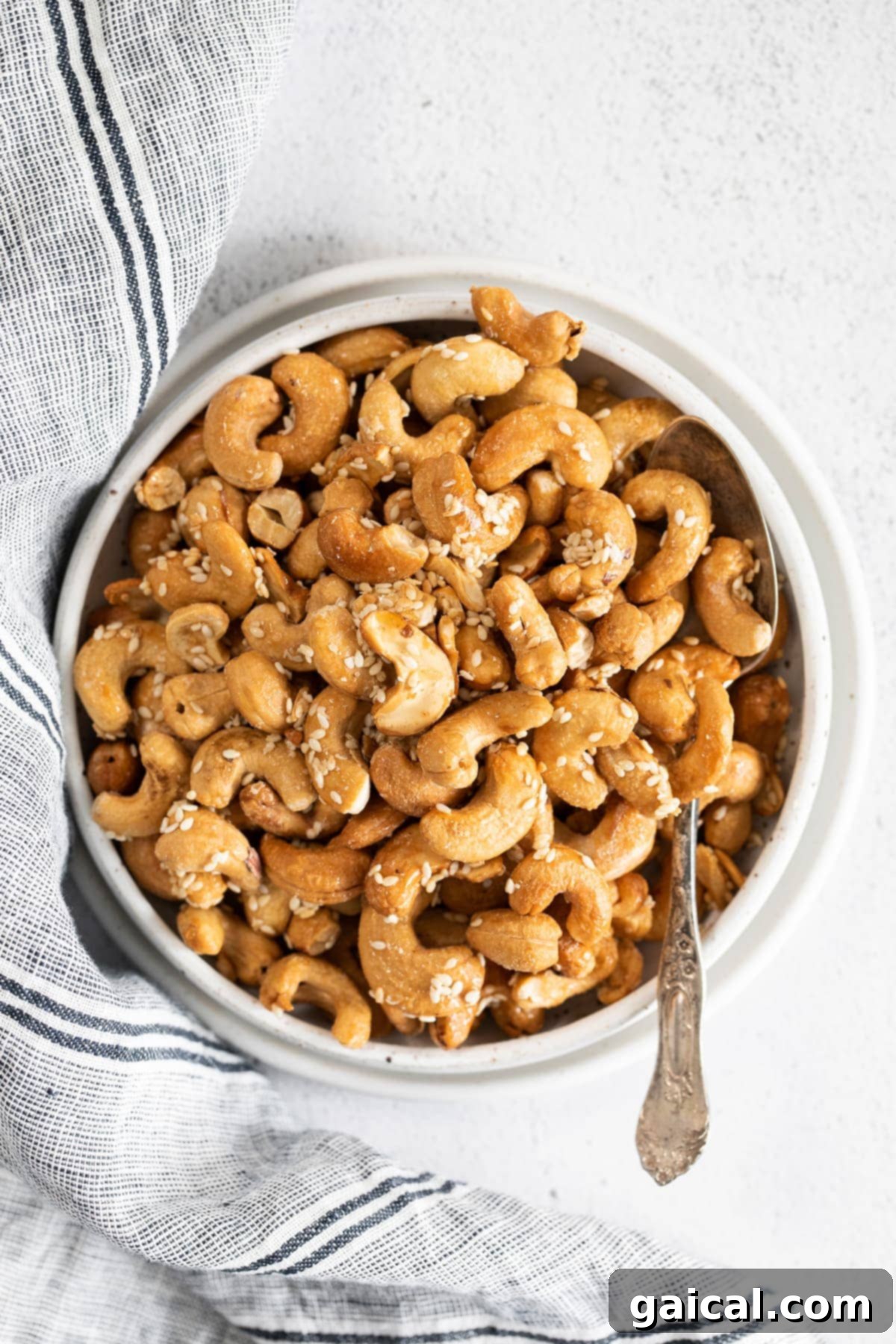 A close-up shot of roasted cashews with sesame seeds in a small bowl, next to a spoon, highlighting their golden-brown color and appealing texture.