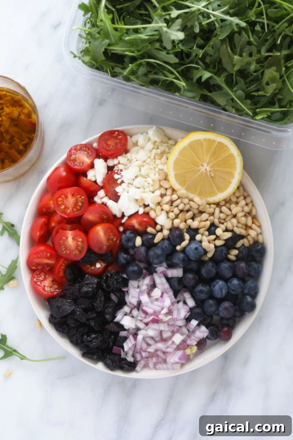 This arugula salad has dried fruit, vegetables, cheese, and a delicious dressing! It is the best arugula salad out there. It is perfect for a meal prep lunch or a great addition to any dinner! A beautiful overhead shot showing all the fresh ingredients for the arugula salad neatly arranged on a white plate, showcasing their vibrant colors and textures, ready for assembly.