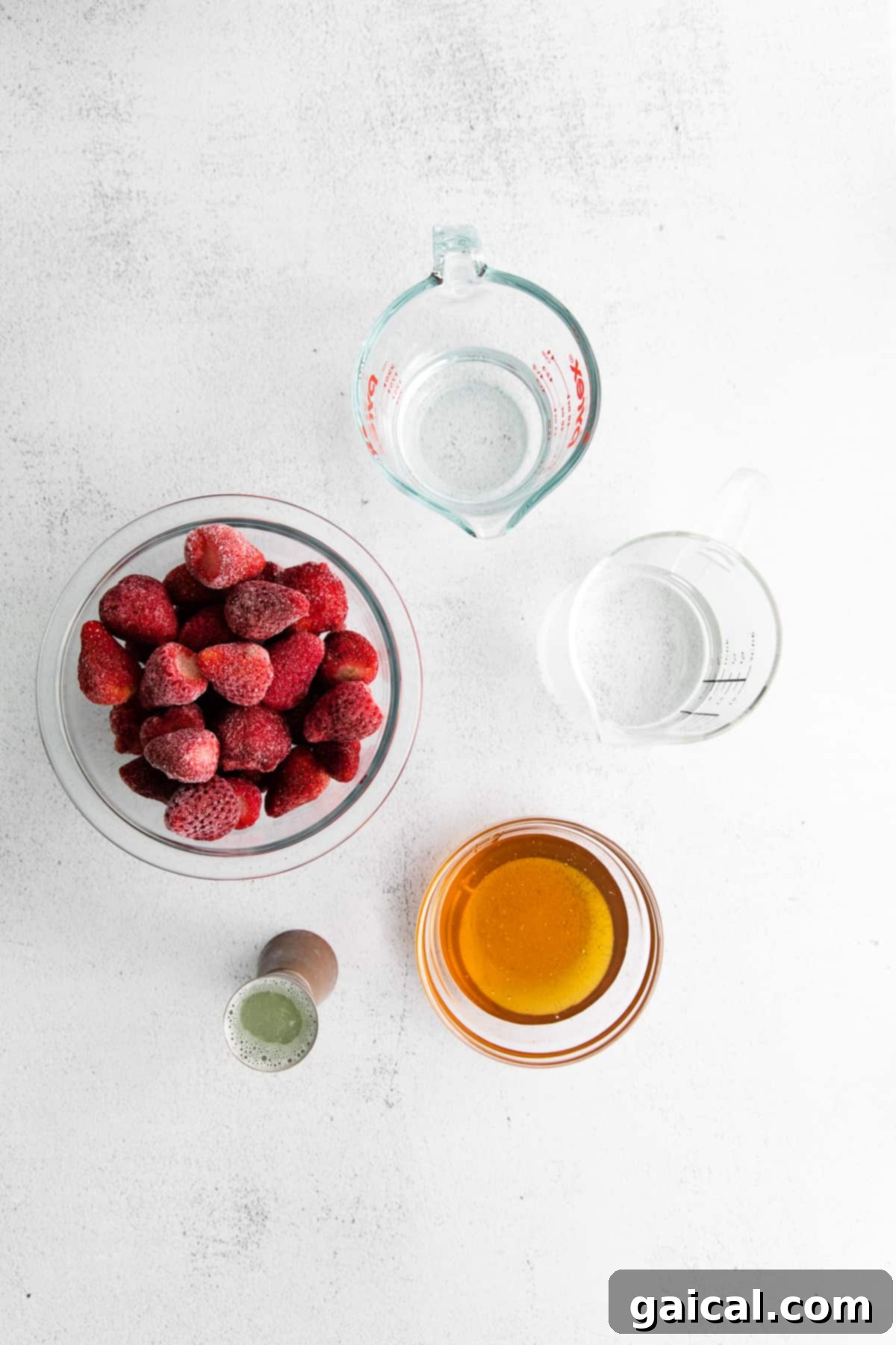 Essential ingredients for a strawberry daiquiri laid out on a kitchen counter: frozen strawberries, rum, limes, and honey simple syrup.