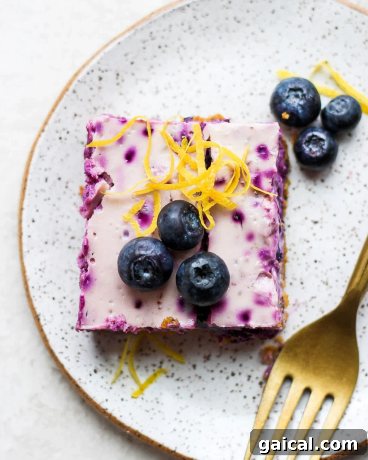 A single blueberry cheesecake bar on a white plate, garnished with fresh blueberries and lemon zest, ready to be enjoyed.