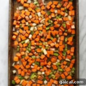 Raw sweet potatoes, bell pepper, and onion, coated in olive oil and spices, spread on a baking sheet before roasting.