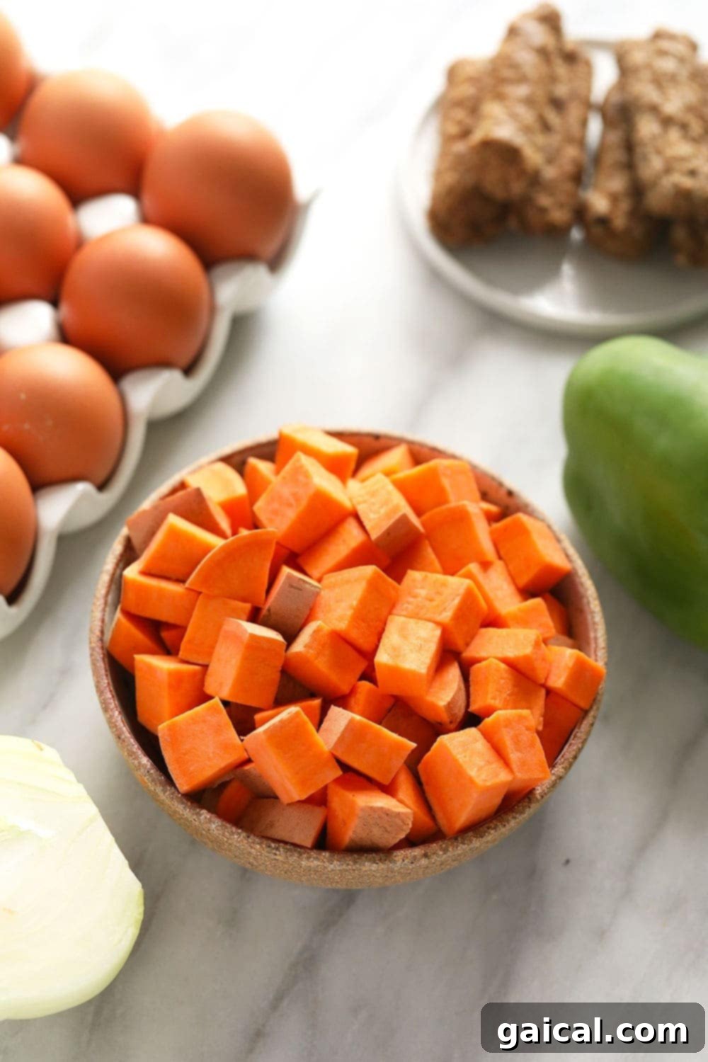 A flat lay of raw ingredients for sweet potato hash: sweet potatoes, eggs, breakfast sausage, a green bell pepper, and an onion, all ready for prep.