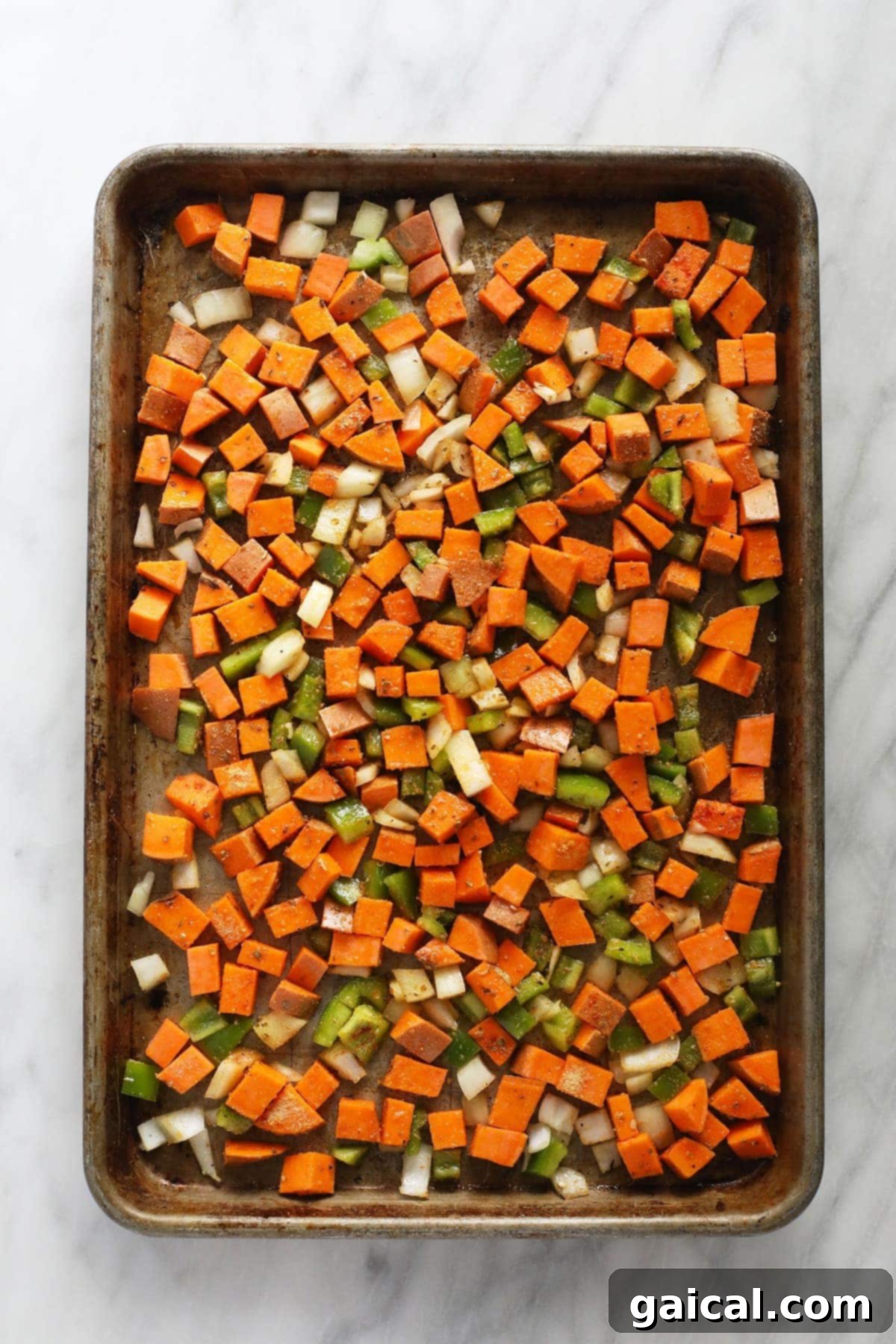 Diced sweet potatoes, bell pepper, and onion spread evenly on a baking sheet, glistening with olive oil and spices, ready for roasting.