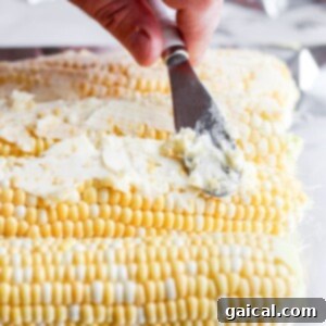 A hand using a spatula to spread garlic butter evenly over an ear of corn.