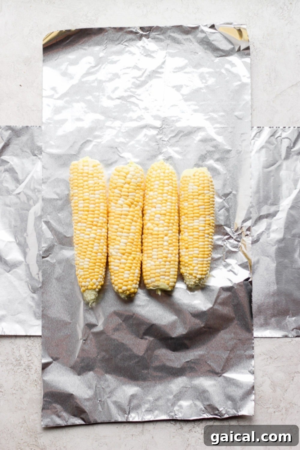 Foil Packet Grilled Corn 3 Four ears of fresh corn on the cob laid out on a sheet of aluminum foil, ready for seasoning and wrapping.