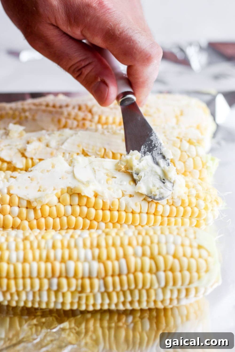 Foil Packet Grilled Corn 4 Softened garlic butter being generously spread on a fresh ear of corn with a spatula, preparing it for grilling in foil.