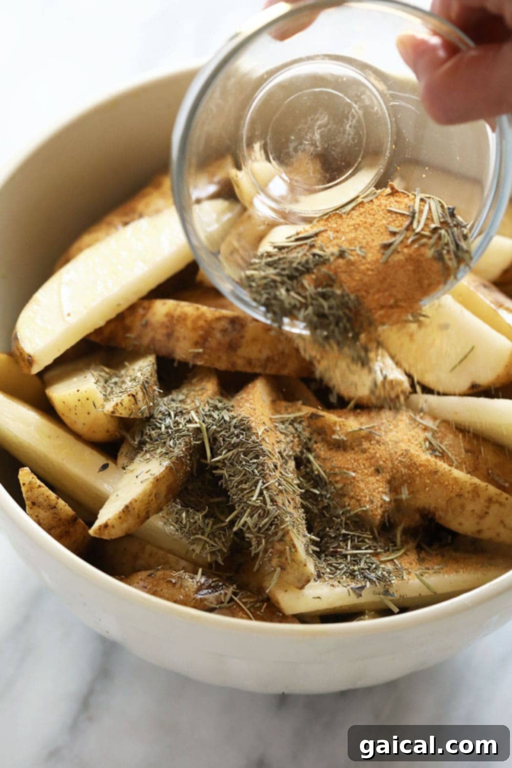 Crispy Roasted Potato Wedges 4 A hand pouring dried herbs and spices over chopped potato wedges in a bowl, ready for tossing.
