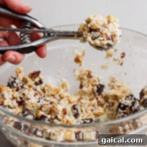 A hand uses a metal cookie scoop to lift dough from a glass bowl filled with a chunky cookie mixture containing nuts and chocolate.