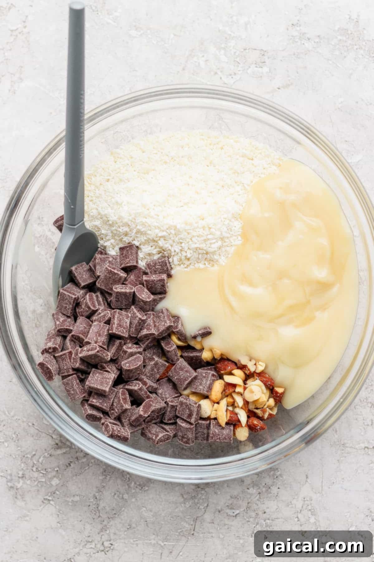 A glass bowl containing sweetened condensed milk, chopped nuts, shredded coconut, and chocolate chunks with a gray spatula.