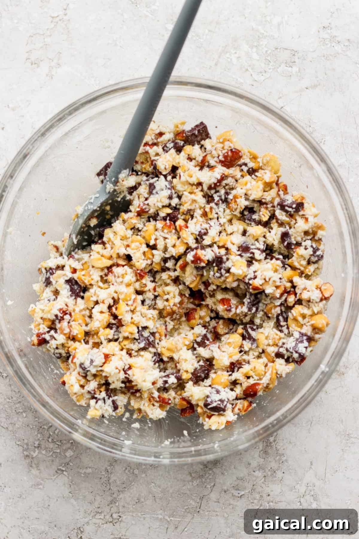 A glass bowl filled with a mixture of chopped nuts, chocolate chunks, and coconut flakes, with a gray spatula resting inside.