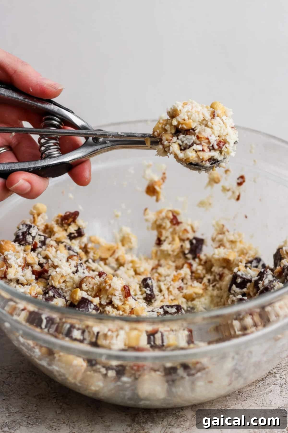 A hand uses a metal cookie scoop to lift dough from a glass bowl filled with a chunky cookie mixture containing nuts and chocolate.