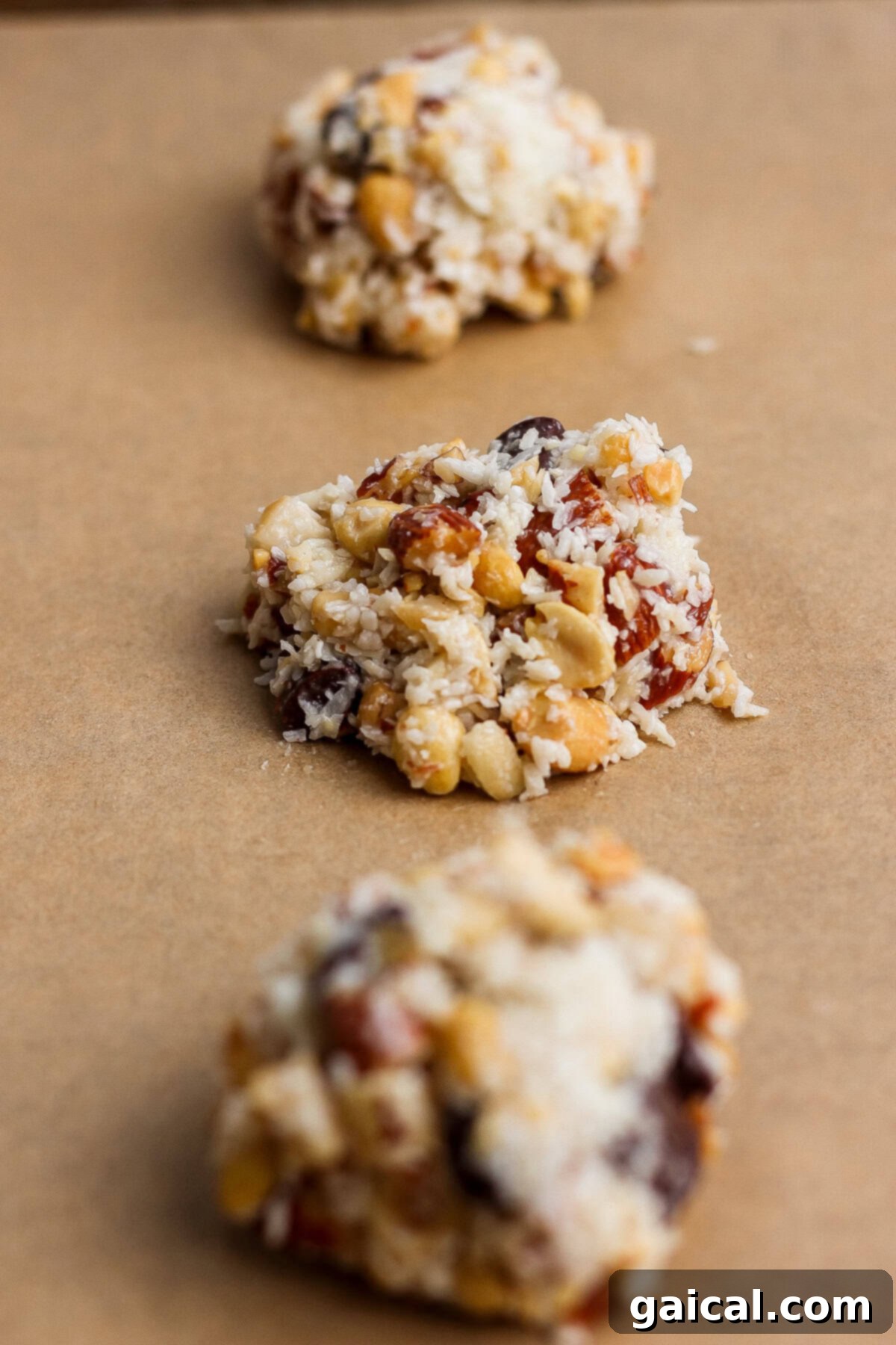 Three unbaked clusters of mixed nuts, dried fruit, and shredded coconut on a sheet of brown parchment paper.