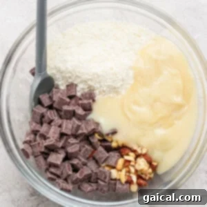 A glass bowl containing sweetened condensed milk, chopped nuts, shredded coconut, and chocolate chunks with a gray spatula.