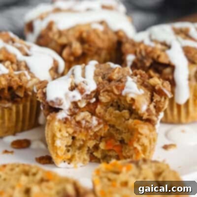Close-up of a carrot muffin with a crumbly topping and white icing. One muffin has a bite taken out, revealing a moist interior with visible carrot pieces.