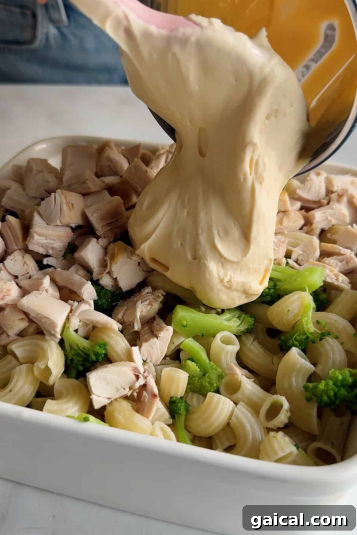 A luscious cheese sauce is being generously poured over a medley of chopped chicken, broccoli florets, and cooked pasta, all nestled in a pristine white baking dish, promising a delightful culinary experience.