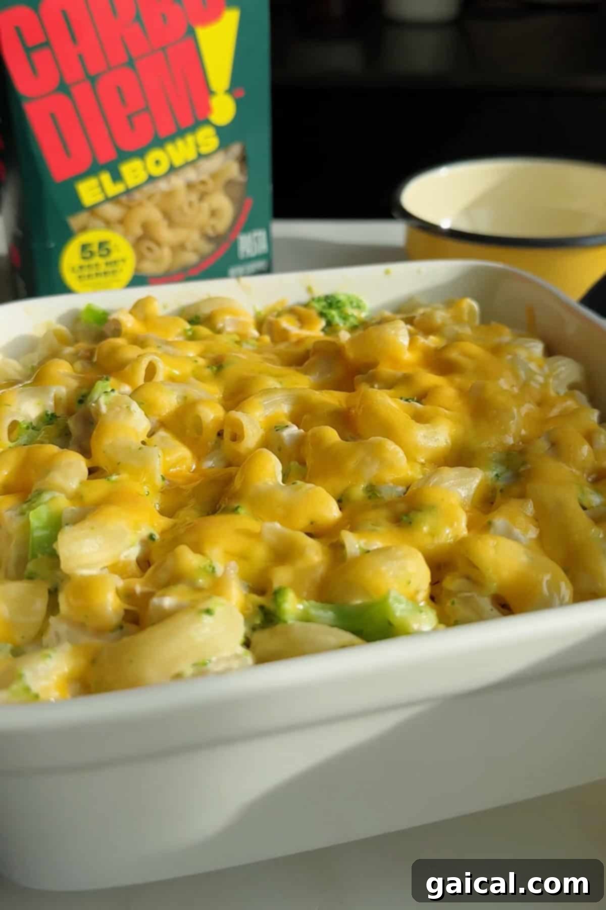 A white dish brimming with creamy macaroni and cheese, adorned with melted cheese and tender broccoli pieces; a box of elbow pasta and a measuring cup subtly placed in the background, enhancing the scene's culinary appeal.