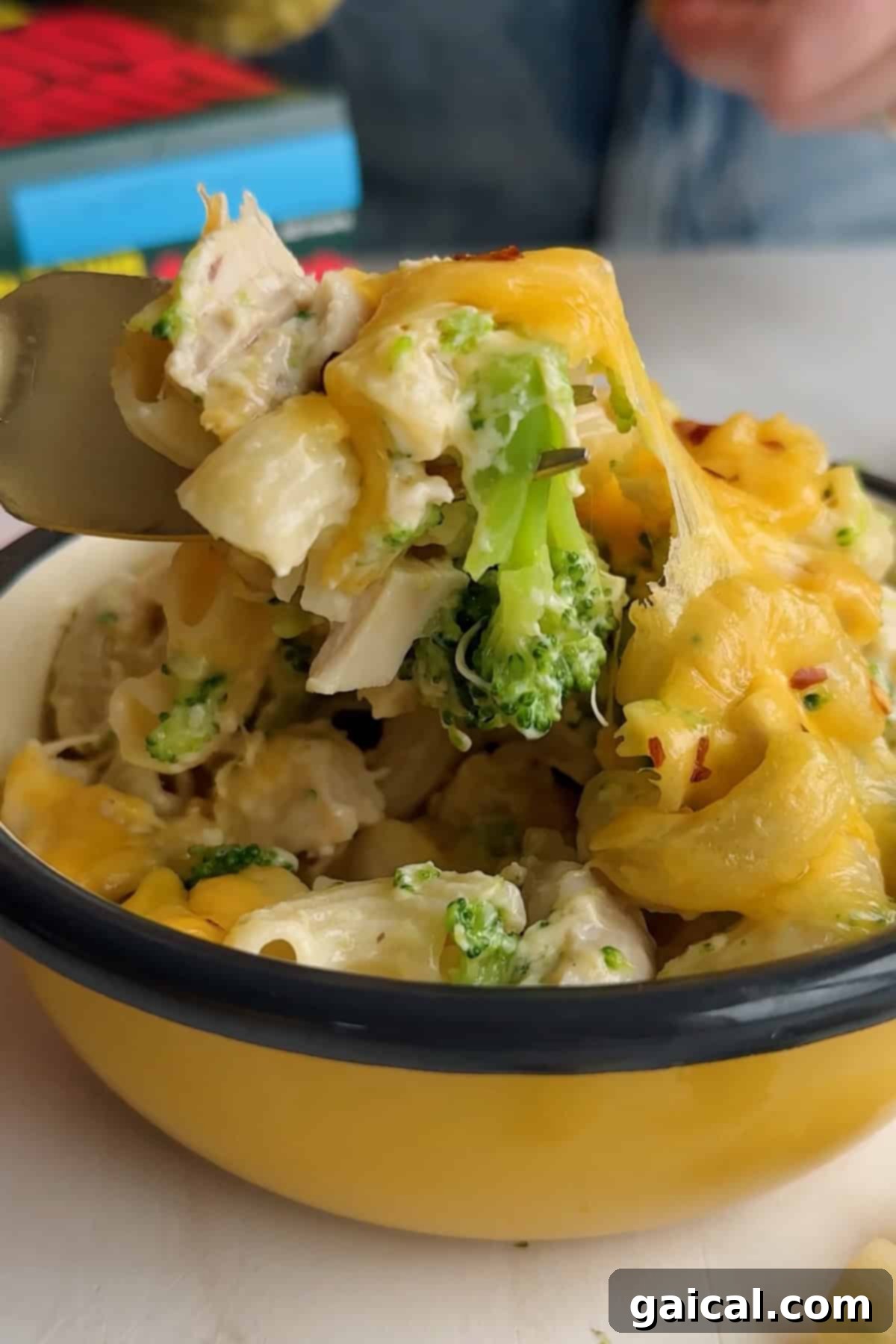 A fork delicately lifts a portion of cheesy pasta, complete with broccoli and chicken, from a vibrant yellow enamel bowl, showcasing the dish's appealing texture and vibrant colors.