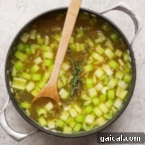 A pot of vegetable soup with chopped celery and herbs, stirred with a wooden spoon, sits on a light-colored surface.