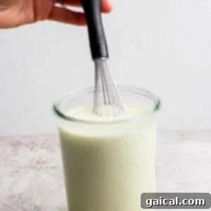 A hand holding a whisk over a glass jar filled with a creamy, pale liquid on a light-colored surface.