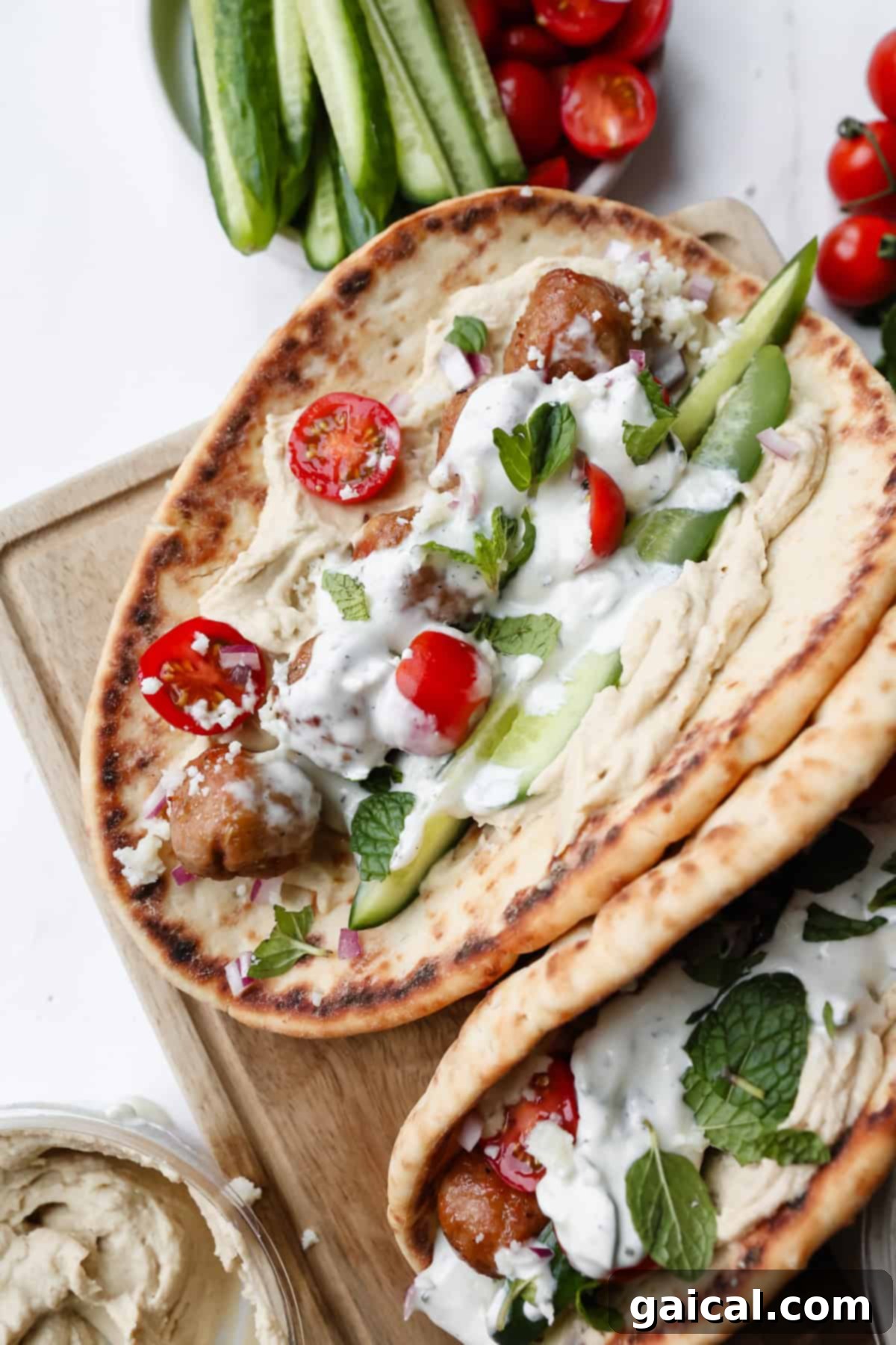 Two flatbreads topped with meatballs, hummus, sliced cucumbers, cherry tomatoes, tzatziki sauce, fresh herbs, and feta cheese on a wooden board.
