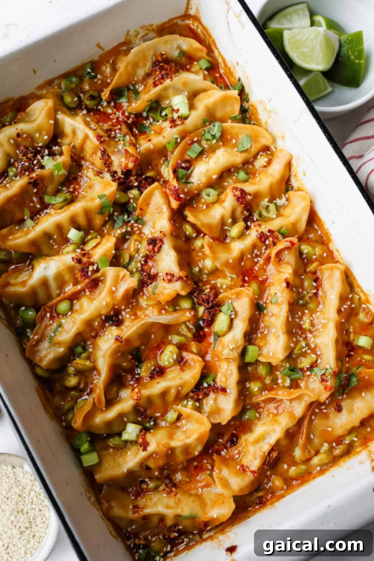 A white baking dish filled with cooked dumplings in a spicy sauce, garnished with chopped green onions, cilantro, and sesame seeds. A plate of lime wedges is nearby.