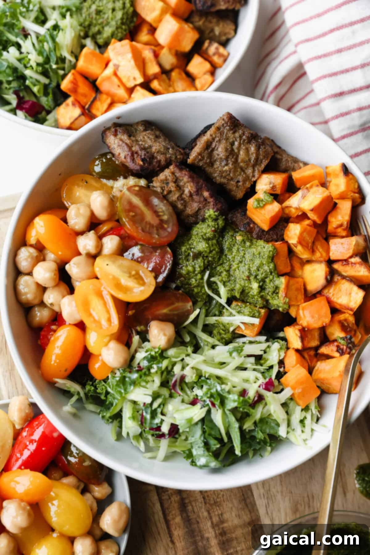 A bowl filled with grilled beef strips, roasted sweet potatoes, cherry tomatoes, chickpeas, shredded greens, and green sauce, with a fork and another bowl partially visible.
