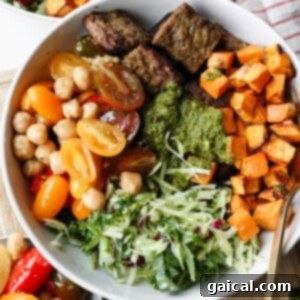 A bowl containing grilled meat, roasted sweet potatoes, cherry tomatoes, chickpeas, shredded greens, and a serving of green sauce.