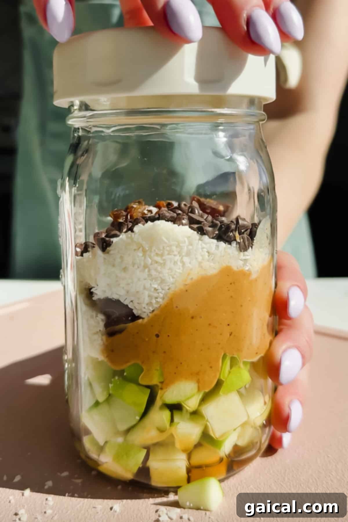 Ingredients for Caramel deLites Apple Salad layered in a glass jar: chopped green apple, peanut butter, shredded coconut, chocolate chips, and cacao nibs.