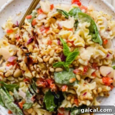 A vibrant plate of pasta salad featuring fresh spinach, toasted pine nuts, hearty white beans, finely chopped red onions, and bell peppers, all drizzled with balsamic glaze and garnished with fresh basil.