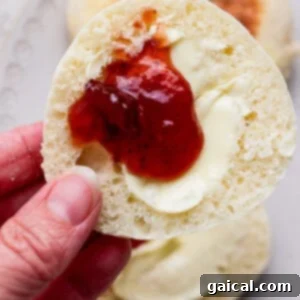 Protein Packed Cottage Cheese English Muffins 15 A hand holds half of an English muffin topped with melted butter and strawberry jam, with more muffins on a plate in the background, ready to be eaten.