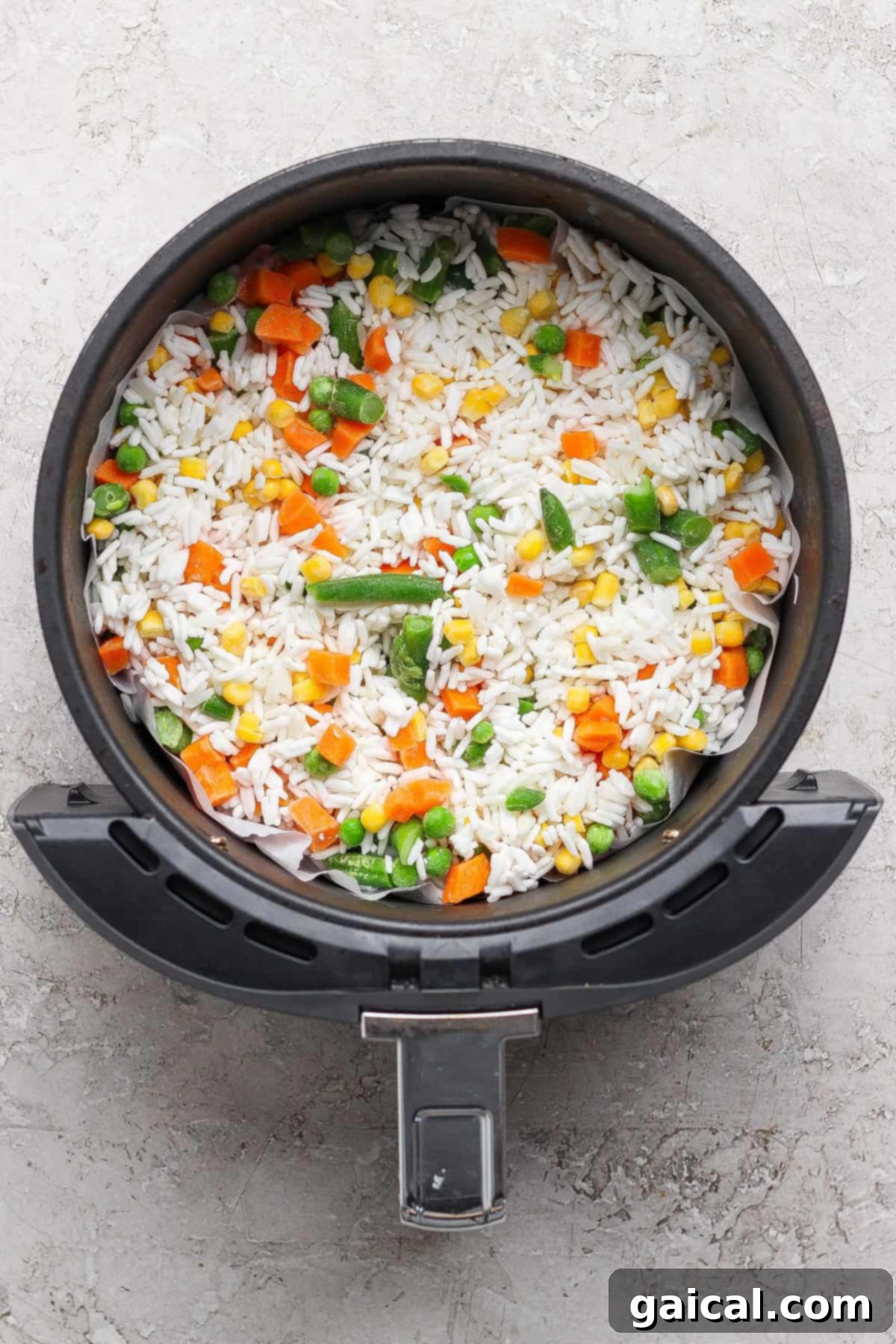 Frozen white rice and a frozen vegetable medley of carrots, peas, and corn are combined in an air fryer basket before cooking.