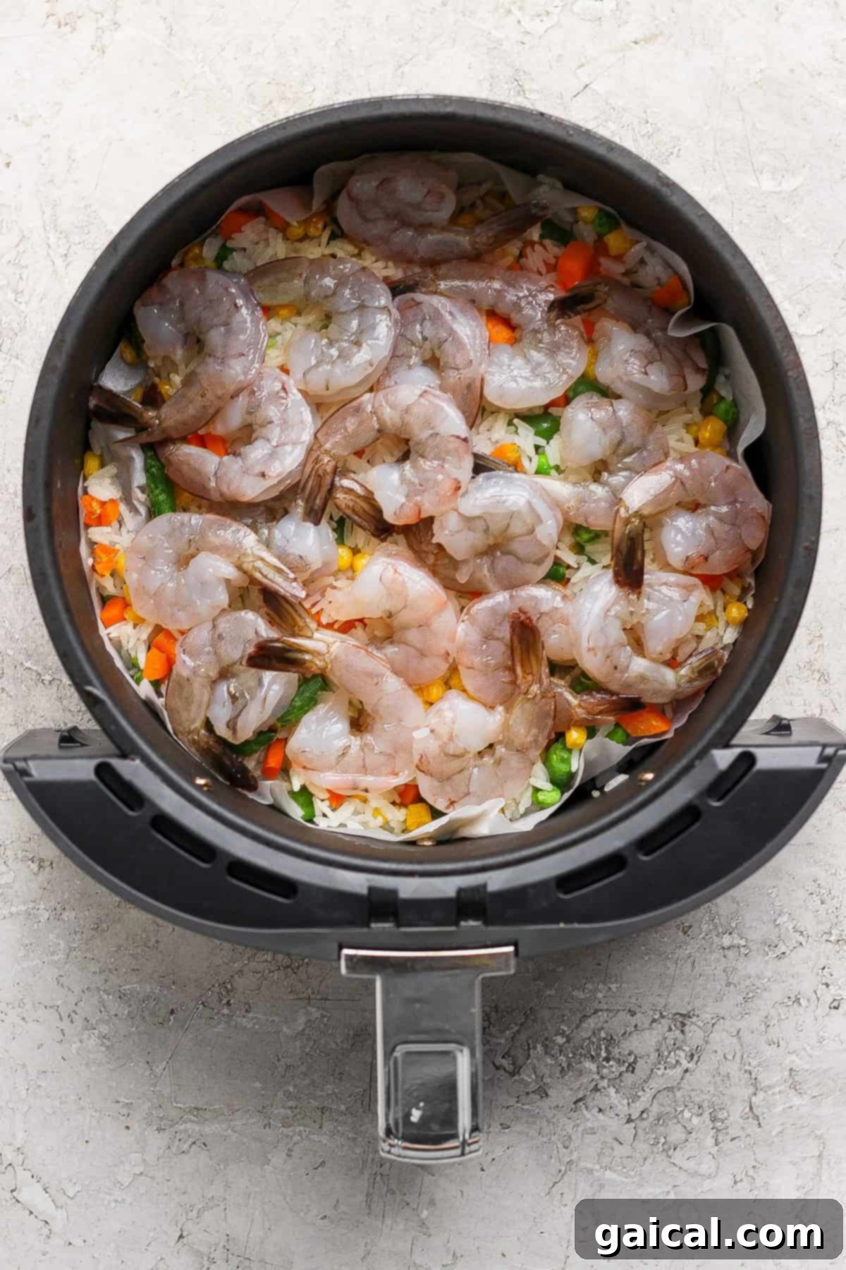 Raw, thawed jumbo shrimp are layered on top of the frozen rice and vegetable mixture inside the air fryer basket, ready for the next cooking stage.