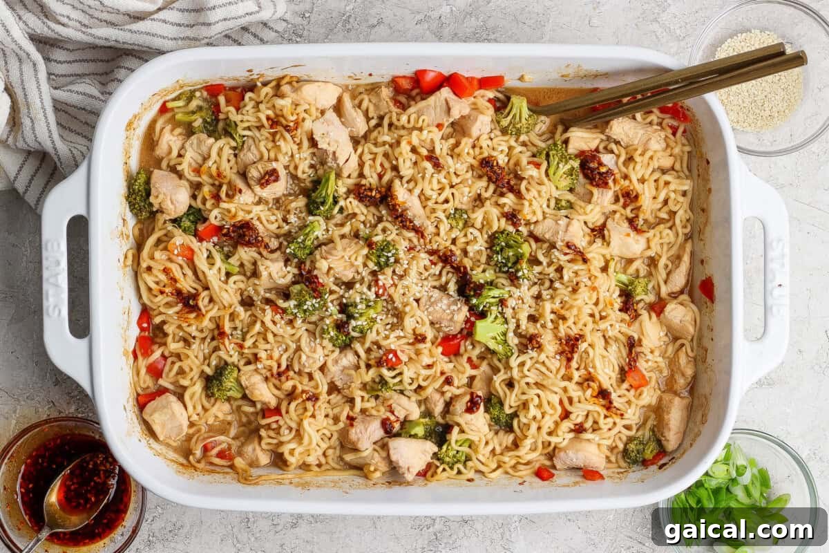 Rectangular white baking dish filled with cooked ramen noodles, chicken pieces, broccoli, and red peppers, garnished with sesame seeds and sauce, with chopsticks resting on the dish.