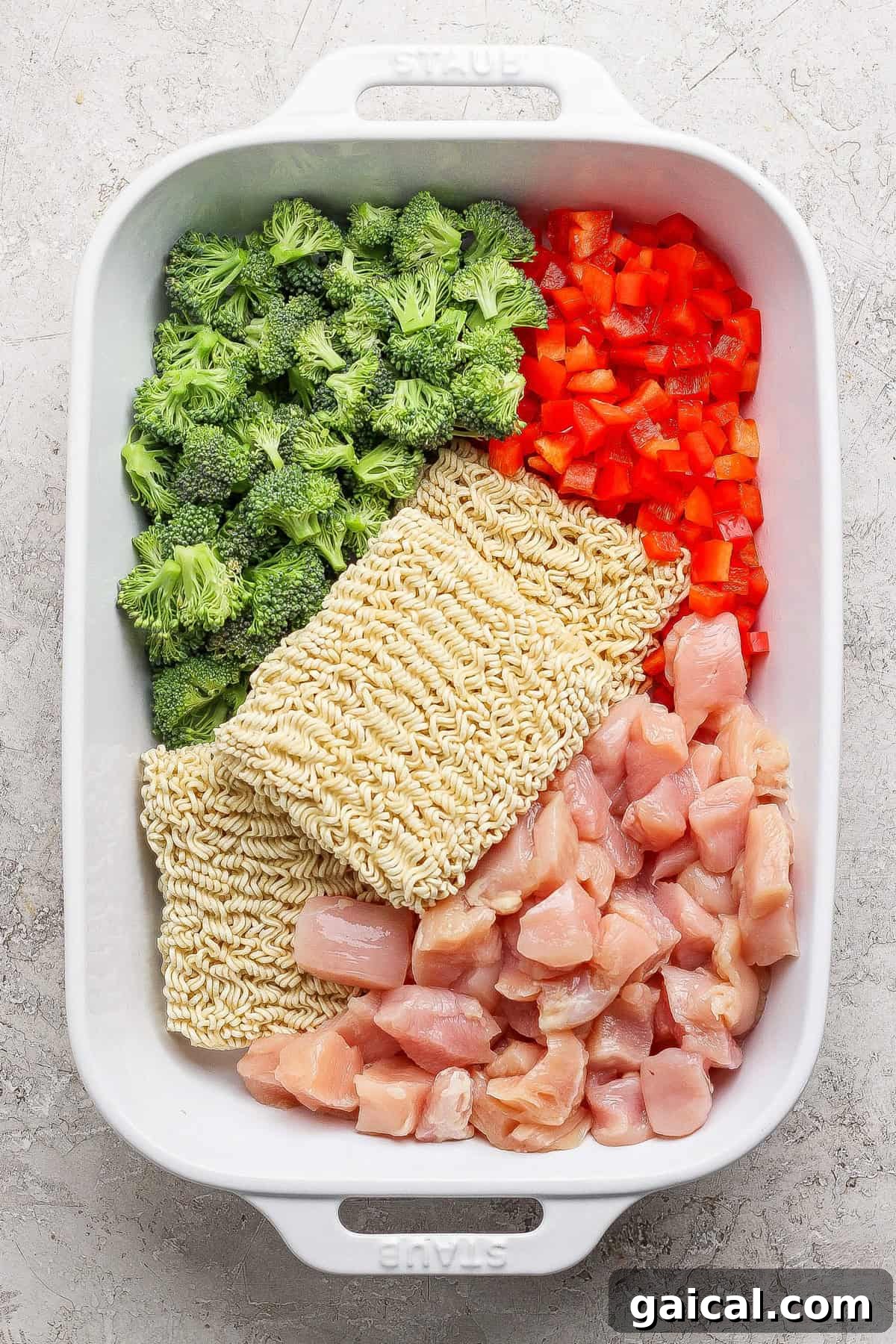 A white baking dish containing raw broccoli florets, diced red bell pepper, two blocks of dry ramen noodles, and chunks of raw chicken, all arranged separately.