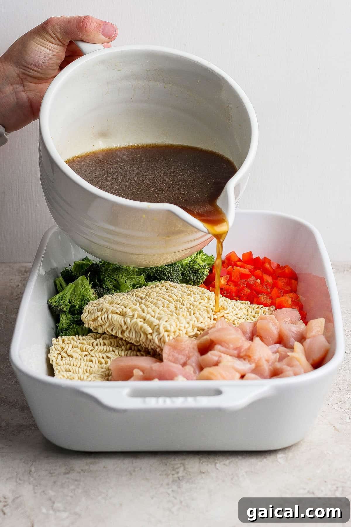 A hand pours brown sauce from a white bowl over uncooked ramen noodles, chopped broccoli, red bell pepper, and diced chicken in a white baking dish.